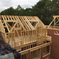 Wooden framing structure of a house under construction, with exposed beams and partially built walls, surrounded by trees.