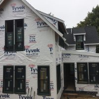 Two-story house under construction, wrapped in Tyvek HomeWrap, with installed windows and partial roofing.