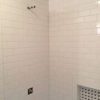 White tiled shower wall with a small shelf, a pipe for a showerhead, and a plumbing fixture for a faucet.