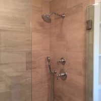 Modern shower with beige tile walls, a round overhead showerhead, handheld showerhead, and metal fixtures.