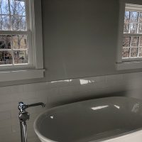 White bathtub beneath two windows with outdoor trees visible, surrounded by white tile walls in a bathroom.