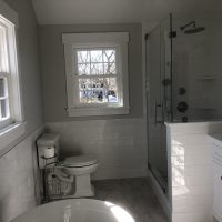 Bright bathroom with white tile, a toilet, glass shower, double windows, and a white vanity under natural light.