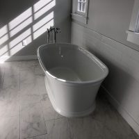 Freestanding white bathtub in a bright bathroom with tile flooring and sunlight streaming through a window.